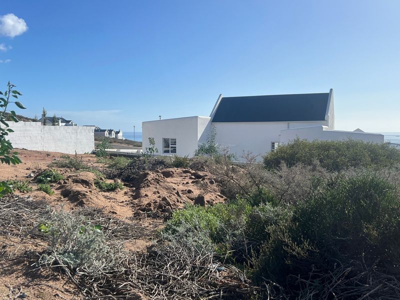 A view of a vacant residential plot with natural coastal vegetation, showing adjacent white-walled properties and a glimpse of the ocean.