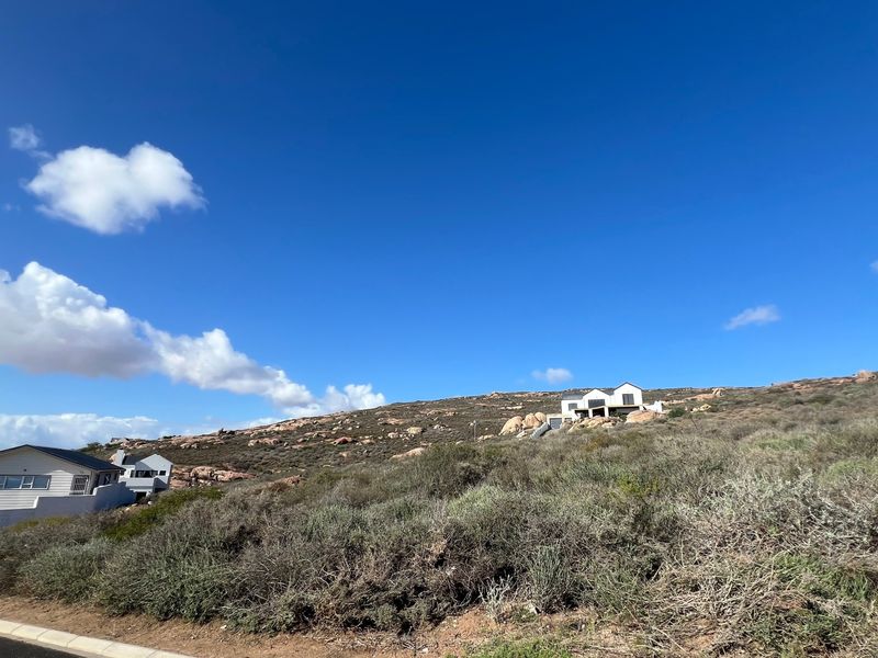 A wide-angle view of a sloped residential vacant land plot featuring natural coastal vegetation and surrounding hillside homes.
