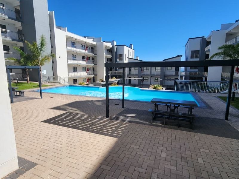 A well-maintained communal swimming pool area featuring paved patio space, pergola structures, and landscaped surroundings within an apartment complex.