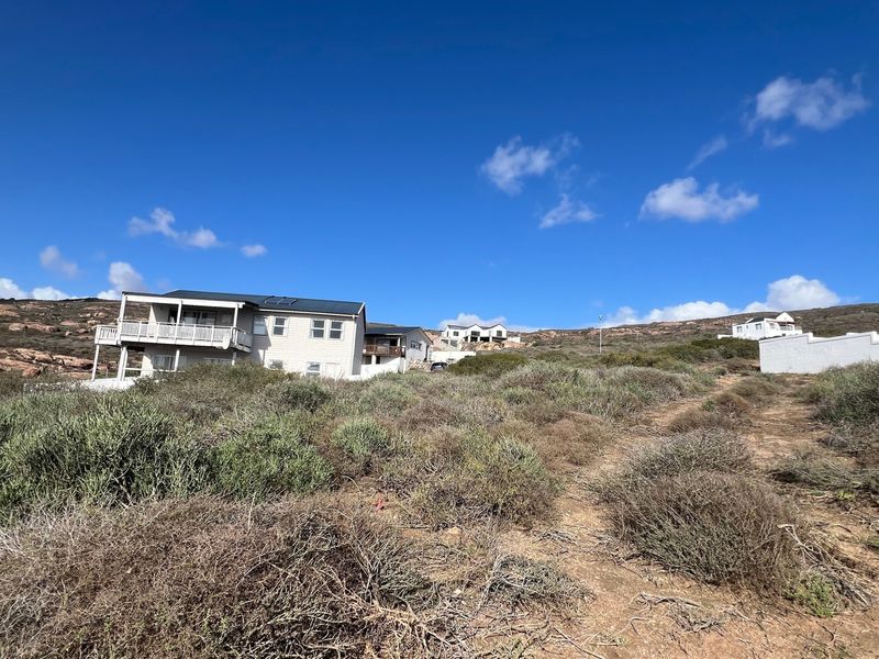 A view of a vacant residential plot featuring natural coastal vegetation with established neighboring properties visible in the background.