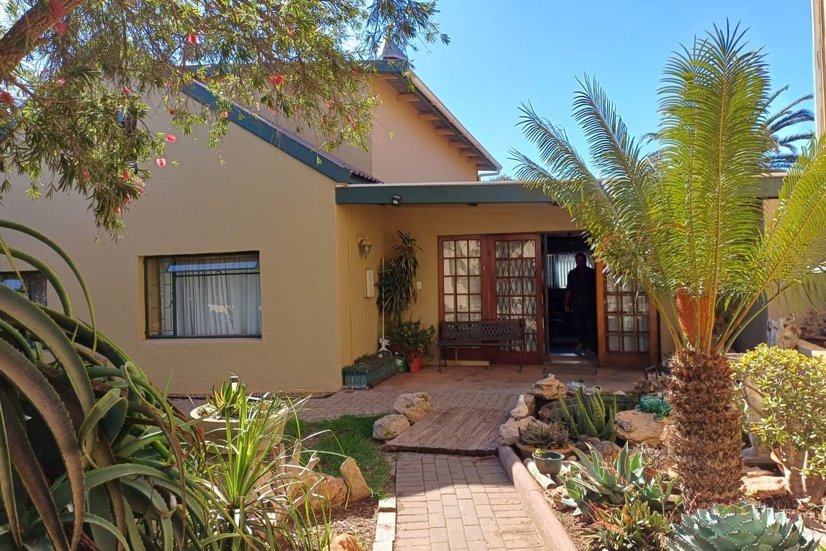 Rear view of the property featuring a covered patio, brick paved pathway, and established garden with mature landscaping and timber-framed glass doors.