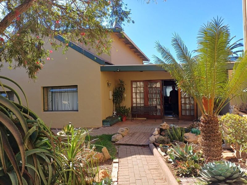 Rear view of the property featuring a covered patio, brick paved pathway, and established garden with mature landscaping and timber-framed glass doors.