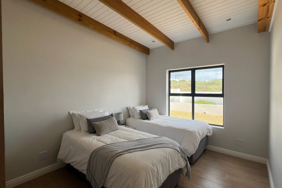 A bright bedroom featuring exposed timber ceiling beams, a white tongue and groove ceiling, laminate flooring, and a large window with an aluminium frame.
