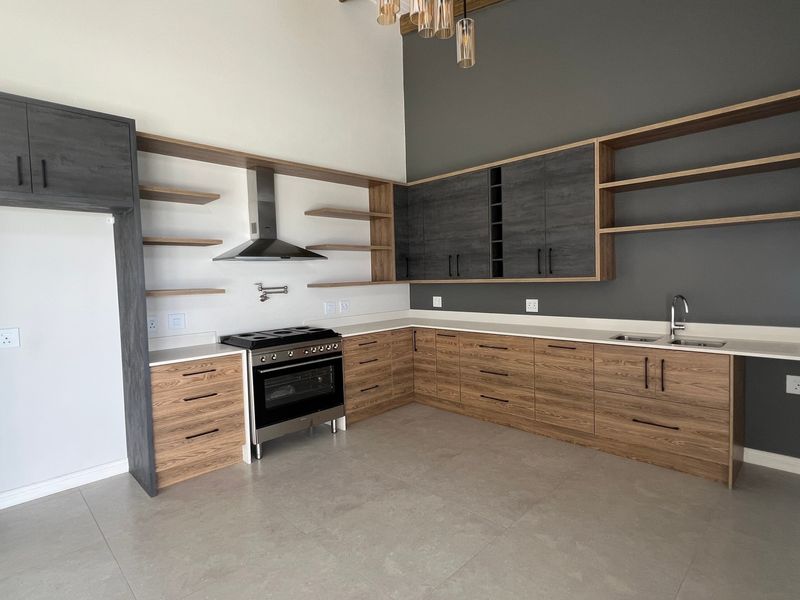 Modern kitchen featuring two-tone cabinetry, stone countertops, stainless steel stove with extractor, double sink, and open shelving in a spacious, light-filled layout.