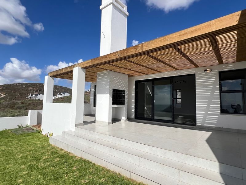 A modern covered patio featuring a timber pergola, built-in braai, and large sliding glass doors, overlooking a landscaped garden area.