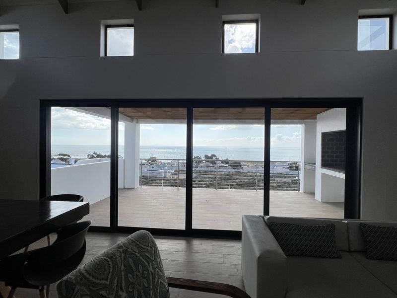 Spacious open-plan living area featuring high ceilings with exposed rafters, large sliding doors opening to a balcony with ocean views, and a built-in braai.