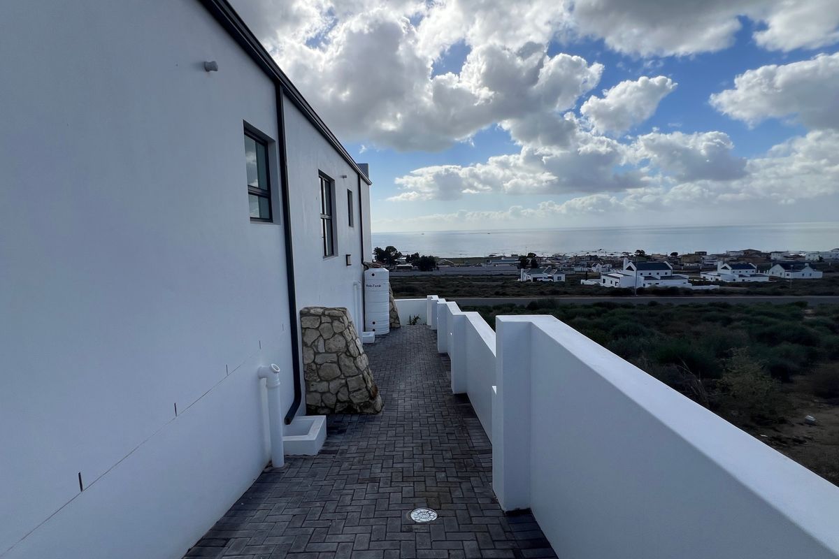 A clean, modern exterior rear walkway featuring stone-clad accents, a water tank, and a parapet wall overlooking a coastal landscape.