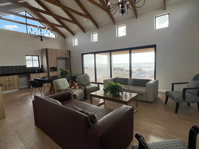 Spacious open-plan living and kitchen area featuring vaulted timber beam ceilings, modern cabinetry, and large sliding doors opening to a scenic coastal view.
