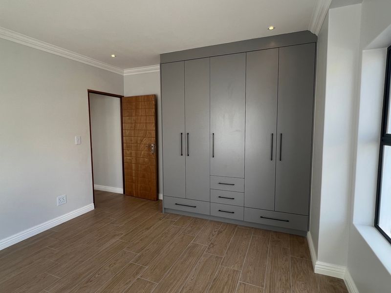 A bright, modern bedroom featuring neutral walls, wood-look tiled flooring, and extensive grey built-in cabinetry with contemporary hardware.