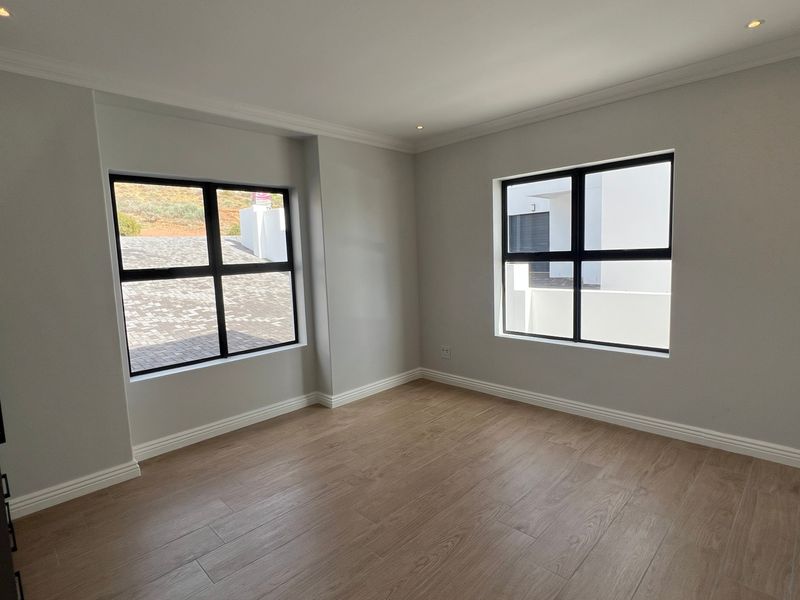 A bright, empty bedroom featuring modern wood-look tile flooring, neutral walls, recessed lighting, and black aluminium-framed windows.