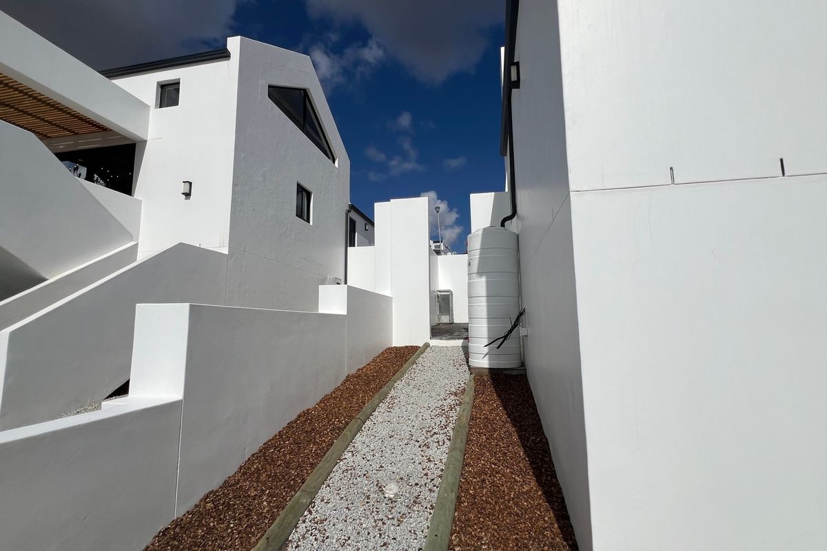 A clean, modern exterior side passage featuring white rendered walls, a gravel pathway with timber edging, and a functional water storage tank.