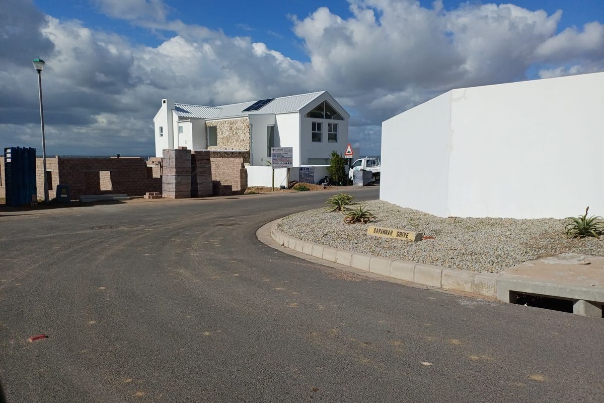 Street view of a residential development area featuring paved roads, concrete curbing, and established boundary walls with a house under construction in the background.