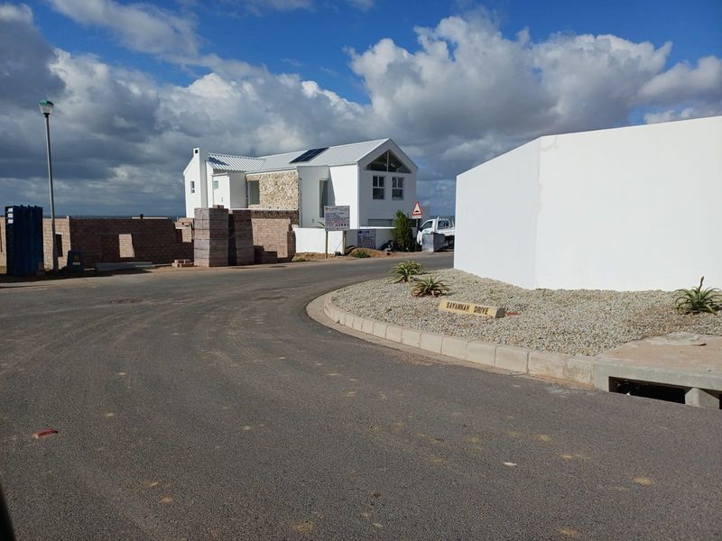 Street view of a residential development area featuring paved roads, concrete curbing, and established boundary walls with a house under construction in the background.