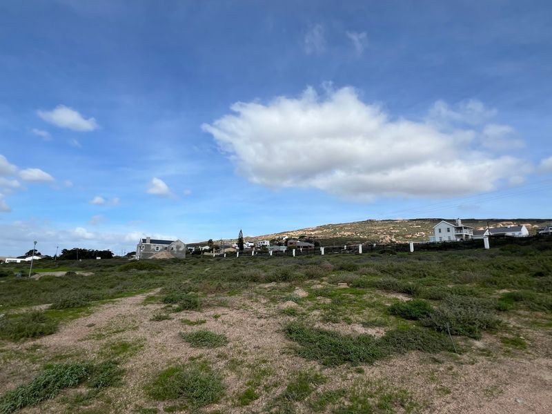 A wide-angle view of a vacant residential plot featuring natural vegetation and perimeter fencing, situated in a coastal neighborhood setting.