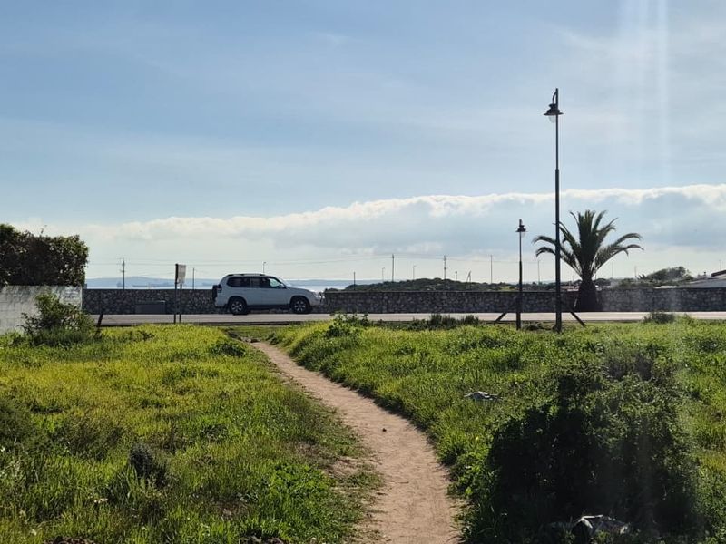 A vacant residential plot featuring an unpaved access path, stone boundary walls, and street lighting, offering a clear view of the adjacent road.