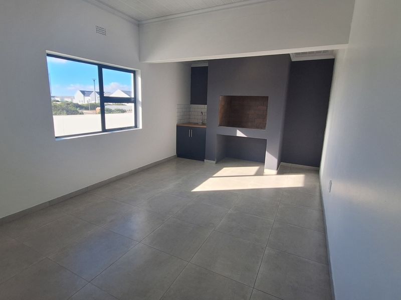 An open-plan living area featuring a built-in braai, kitchenette cabinetry with a sink, and neutral tiled flooring throughout.