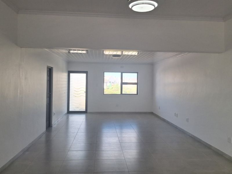 A spacious, empty living area featuring neutral tiled flooring, clean white walls, and natural light from the window and glass-paneled door.