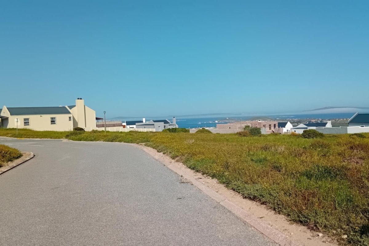 A street-level view of a residential area featuring a paved road, kerbing, and distant coastal views.