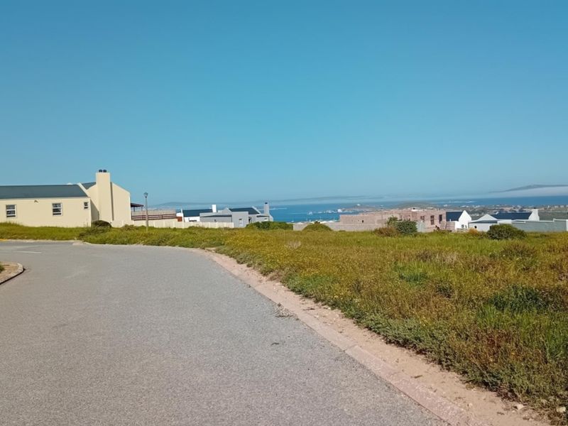 A street-level view of a residential area featuring a paved road, kerbing, and distant coastal views.