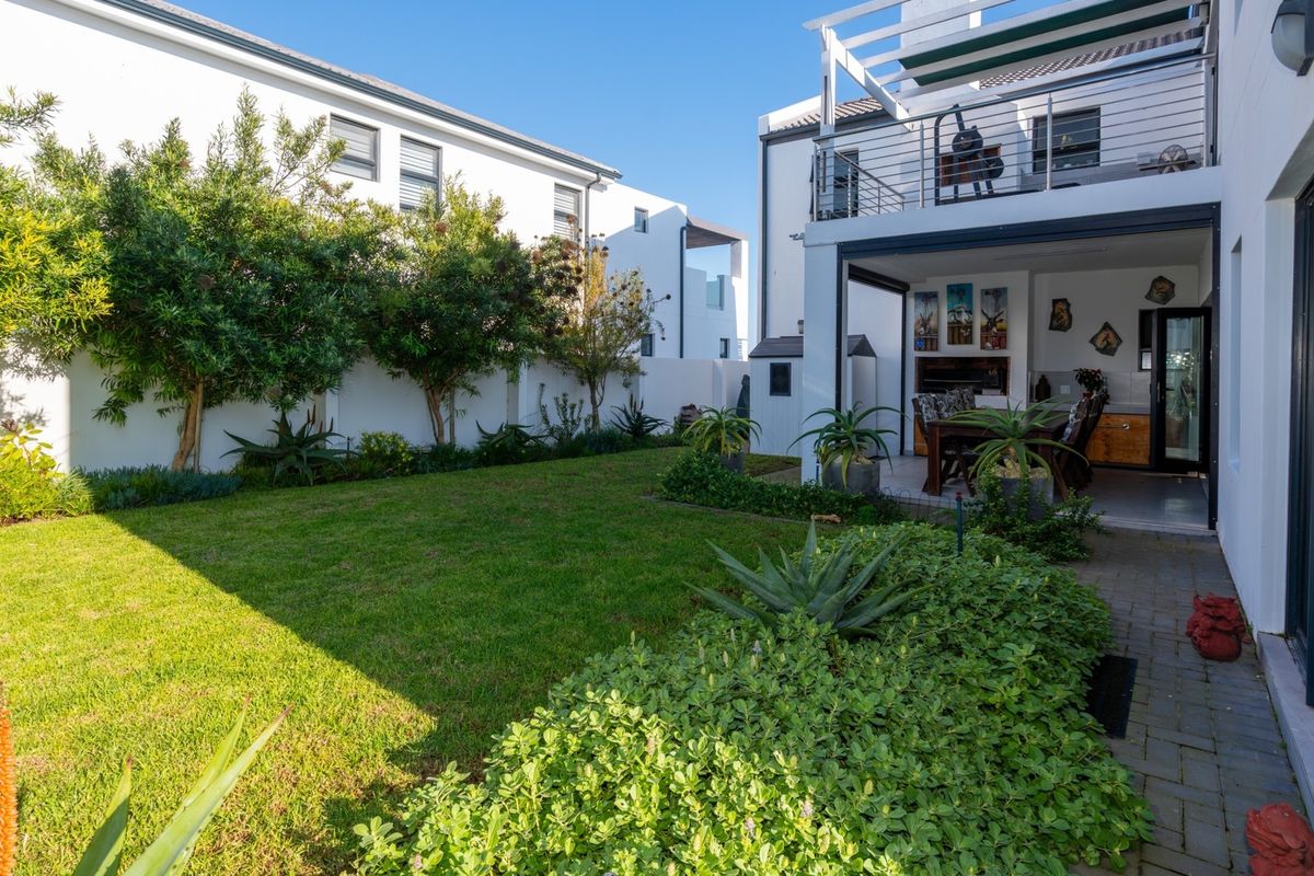 A well-maintained rear garden featuring a lush lawn, landscaped beds, and a covered patio area with a built-in braai and upper balcony.