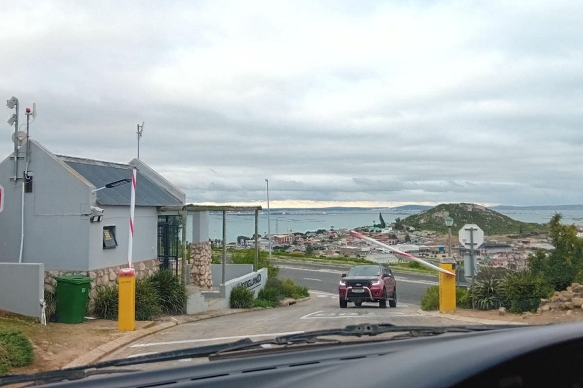 View of the residential estate entrance with a security boom gate, guard house, and surrounding coastal landscape.