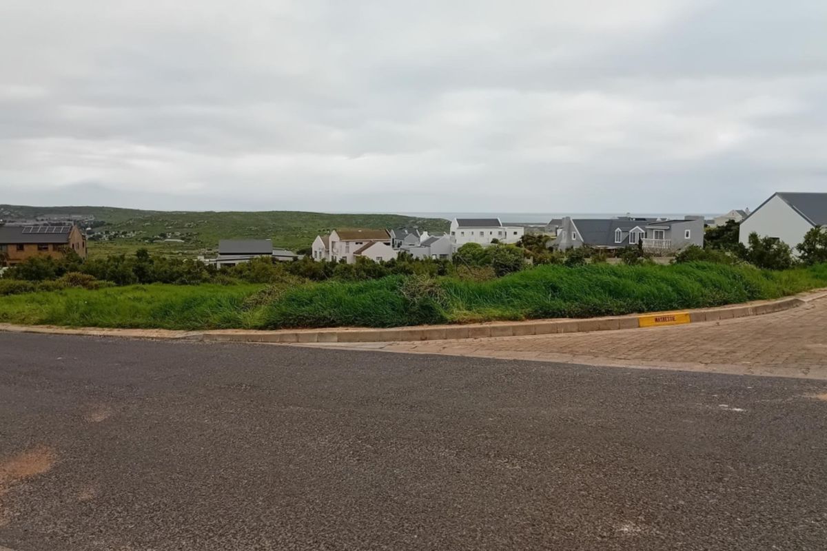 A street-level view of a vacant residential plot featuring paved road access and surrounding coastal neighborhood development.