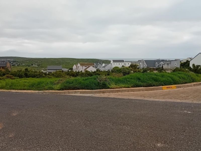A street-level view of a vacant residential plot featuring paved road access and surrounding coastal neighborhood development.