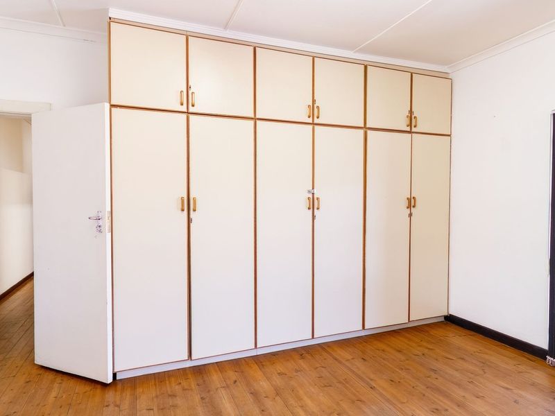 A bright, empty bedroom featuring extensive built-in cupboards, polished wooden flooring, and a secure metal gate over the window.