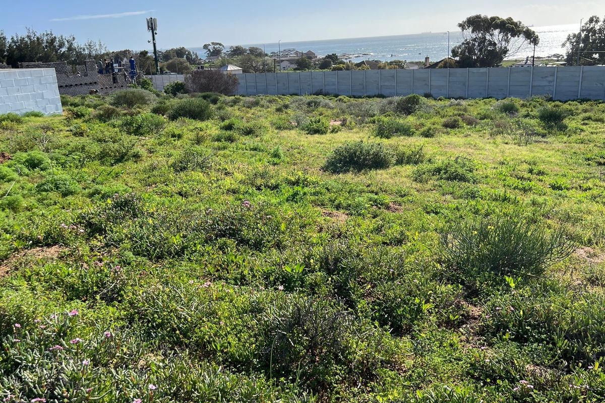 A vacant residential plot featuring natural vegetation, enclosed by a perimeter wall, with distant ocean views and a telecommunications mast visible.