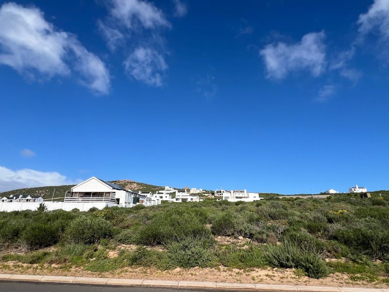 A view of a vacant residential plot featuring native vegetation, situated along a paved road with surrounding coastal residential properties in the background.