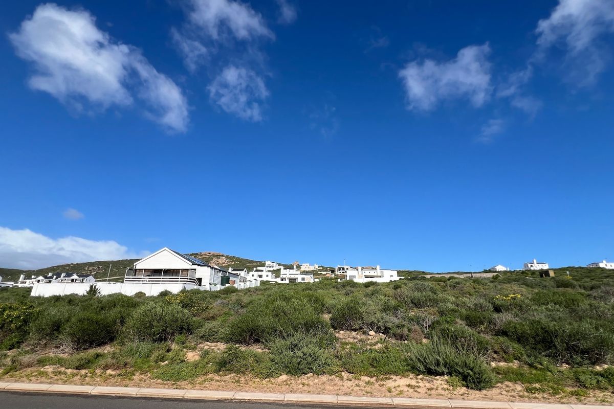 A view of a vacant residential plot featuring native vegetation, situated along a paved road with surrounding coastal residential properties in the background.