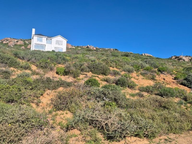 A view of a sloped residential plot featuring natural coastal vegetation and a distant house structure on the hillside.