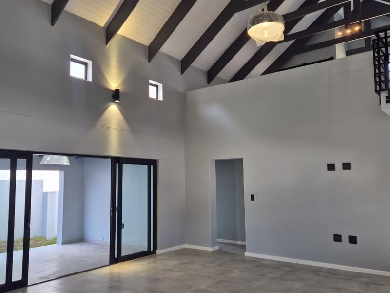 Spacious open-plan living area featuring high vaulted ceilings with exposed rafters, large sliding glass doors, and a mezzanine level with modern metal railings.