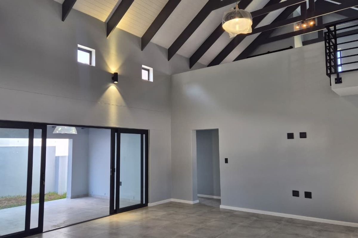 Spacious open-plan living area featuring high vaulted ceilings with exposed rafters, large sliding glass doors, and a mezzanine level with modern metal railings.