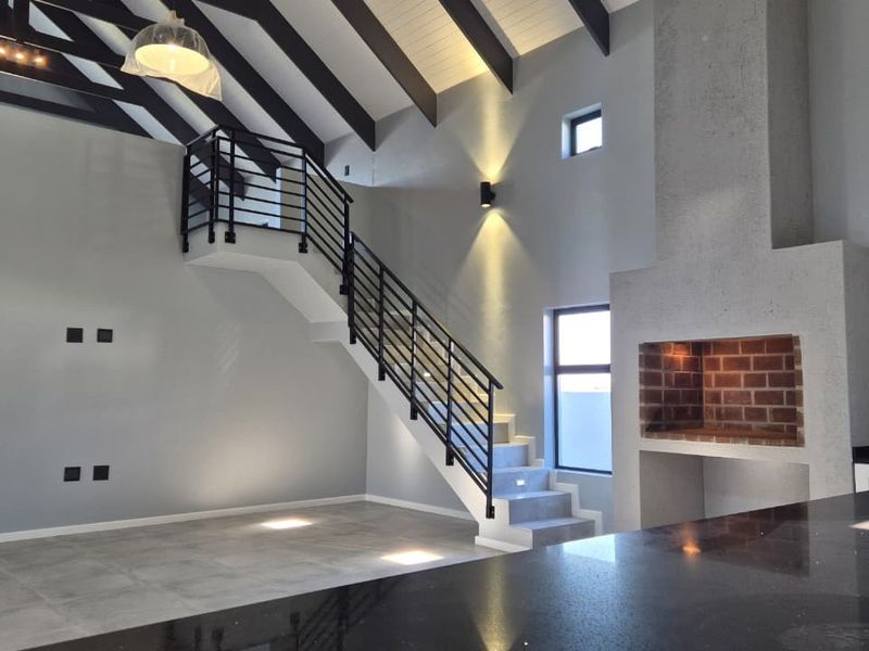 Spacious open-plan living area featuring a vaulted ceiling with exposed beams, a modern staircase, and a built-in fireplace, seamlessly connected to a sleek kitchen.