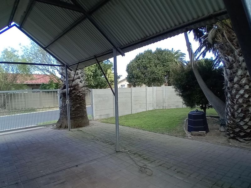 A covered outdoor patio area featuring paved flooring, steel structural supports, and a corrugated metal roof, overlooking a private garden and boundary wall.