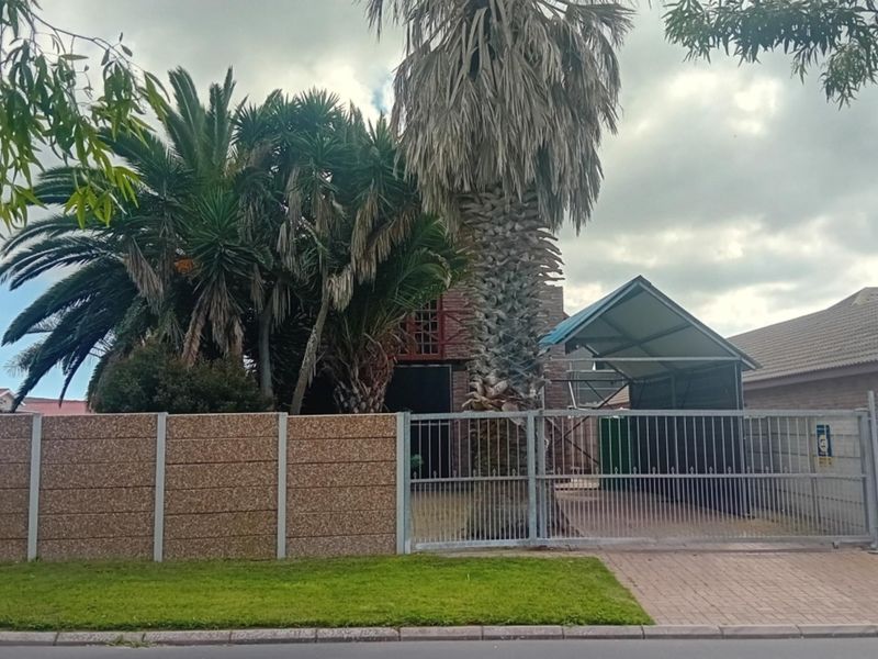Front view of the property featuring a paved driveway, metal gate, carport, and a precast concrete boundary wall with mature palm trees.