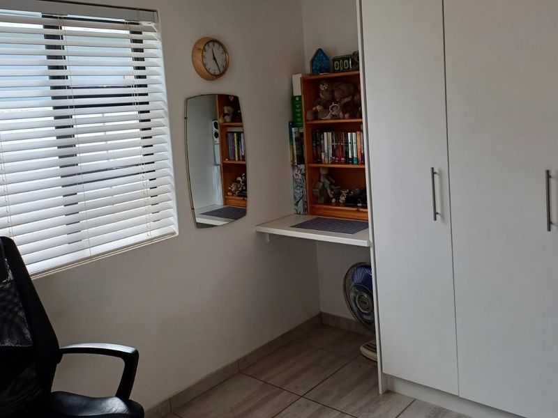 A functional bedroom featuring a built-in wardrobe, a compact built-in desk, and tiled flooring with natural light provided by a window with venetian blinds.