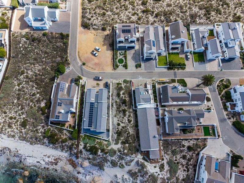 Aerial view of a coastal residential estate featuring modern homes with solar panels, paved driveways, and natural fynbos landscaping near the shoreline.