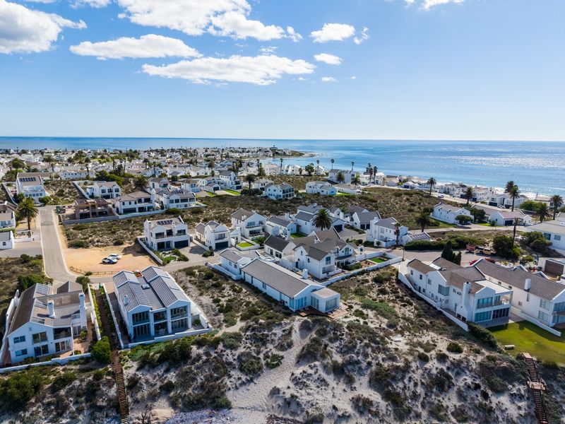 An aerial view of a coastal residential estate featuring white homes, paved streets, and direct access to sandy dunes and the ocean.