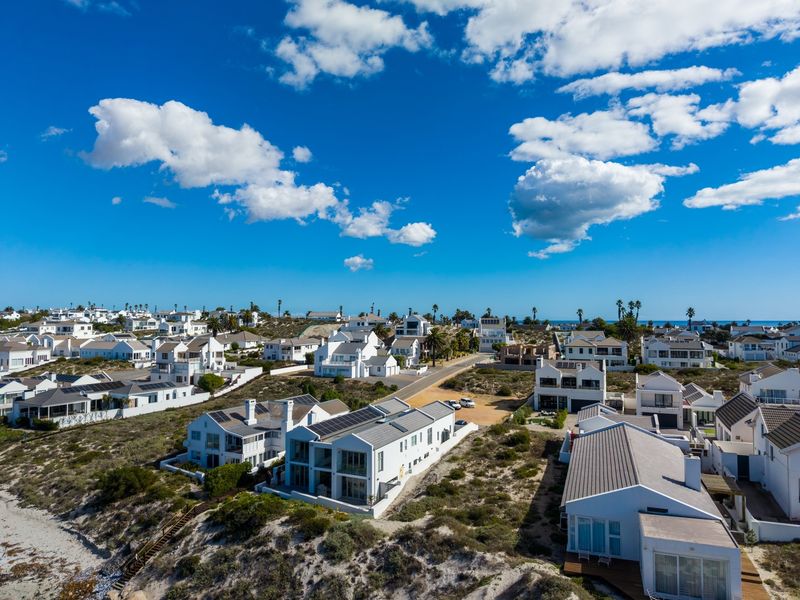 An aerial view of a coastal residential estate featuring white houses, solar installations, and surrounding natural vegetation near the ocean.