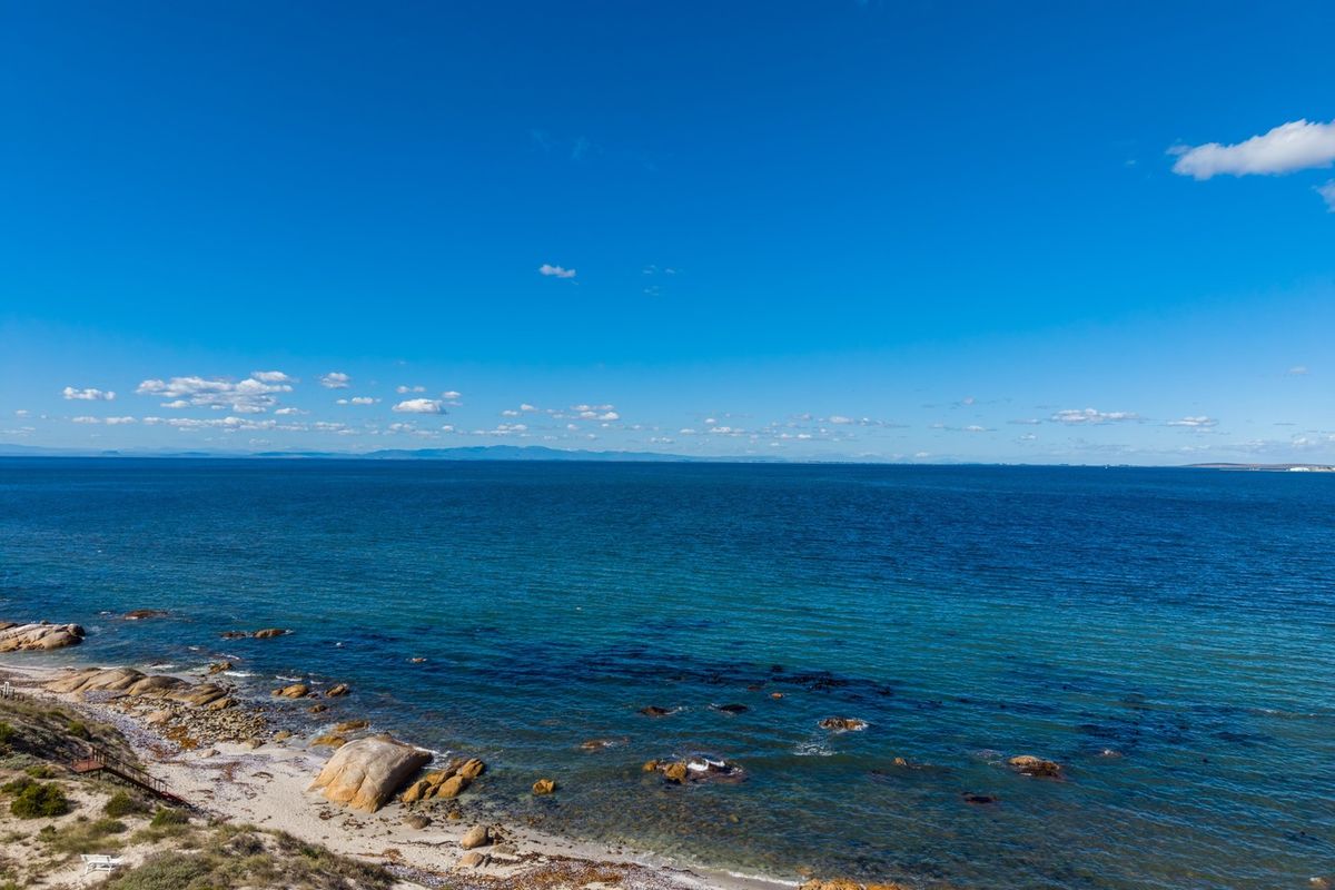 A scenic elevated view of the coastline featuring rocky outcrops, sandy beach, and expansive ocean waters under a clear blue sky.