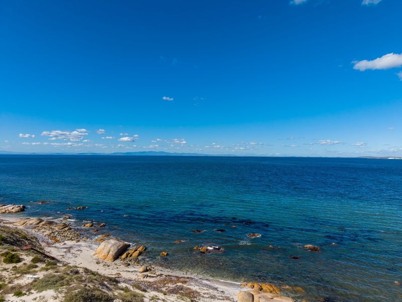 A scenic elevated view of the coastline featuring rocky outcrops, sandy beach, and expansive ocean waters under a clear blue sky.