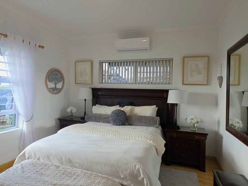 A well-maintained bedroom featuring laminate flooring, a wall-mounted air conditioning unit, and large windows providing natural light and garden views.