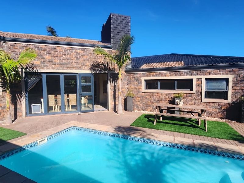 A well-maintained outdoor pool area featuring a paved patio, artificial turf, and a brick-clad rear facade with modern bi-fold doors.