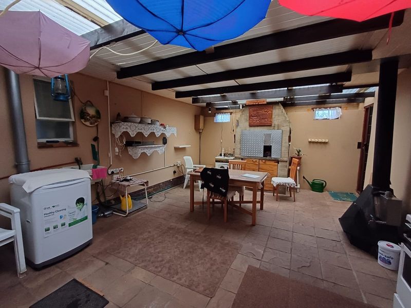 Covered patio area featuring a built-in braai, wood-burning stove, utility sink, and exposed timber beam ceiling with stone tile flooring.