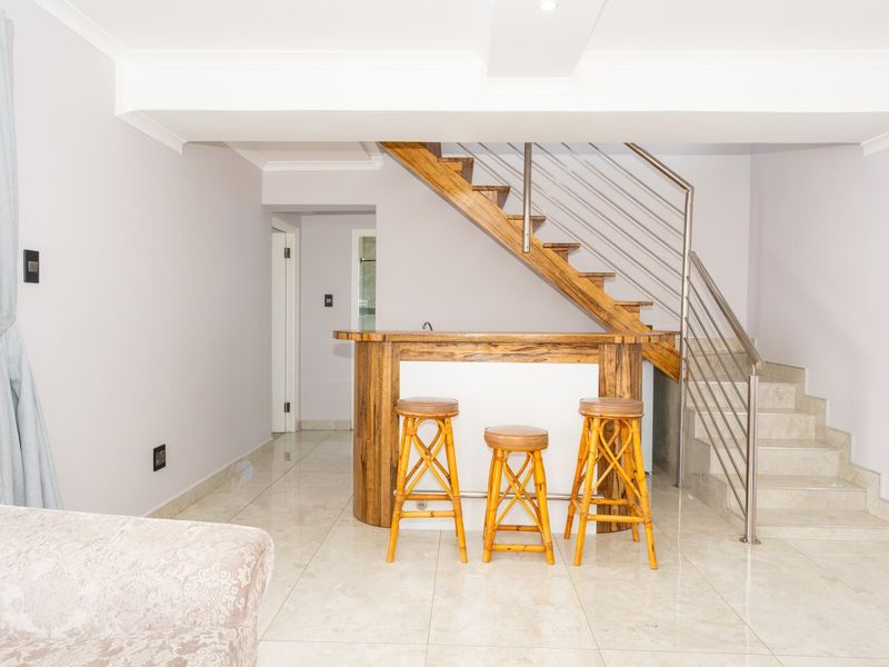 A bright living area featuring a built-in wooden bar, tiled flooring, and a modern staircase with stainless steel balustrades.