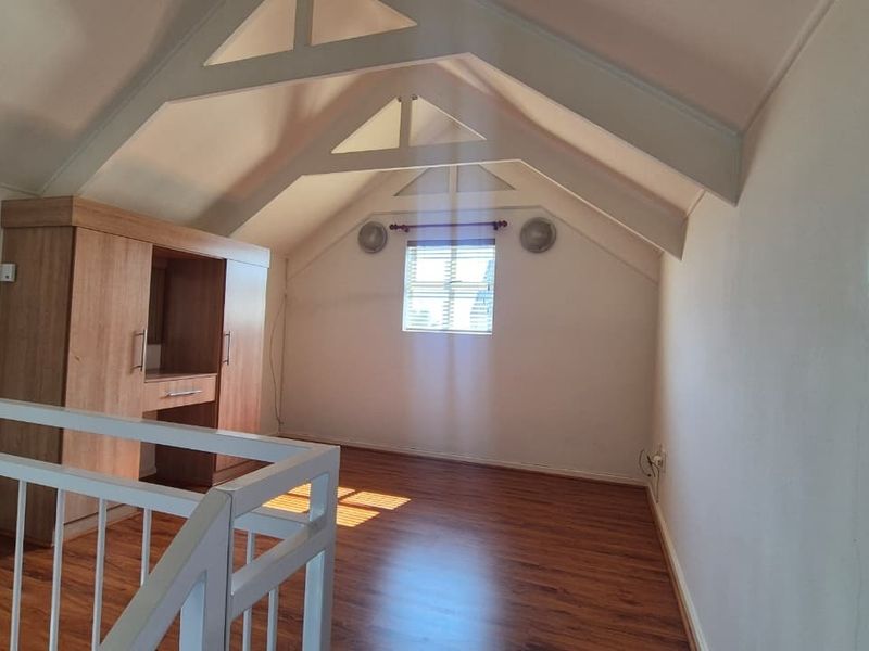 An airy loft-style bedroom featuring vaulted ceilings with exposed trusses, laminate flooring, and built-in cabinetry, overlooking the lower level via a white railing.