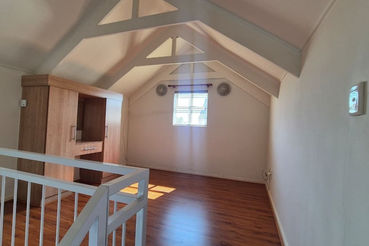 An airy loft-style bedroom featuring vaulted ceilings with exposed trusses, laminate flooring, and built-in cabinetry, overlooking the lower level via a white railing.