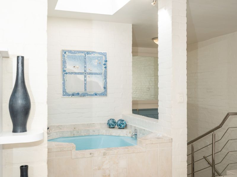 A bathroom featuring a built-in tiled bathtub, exposed white brick walls, and a skylight, adjacent to a staircase with a stainless steel handrail.